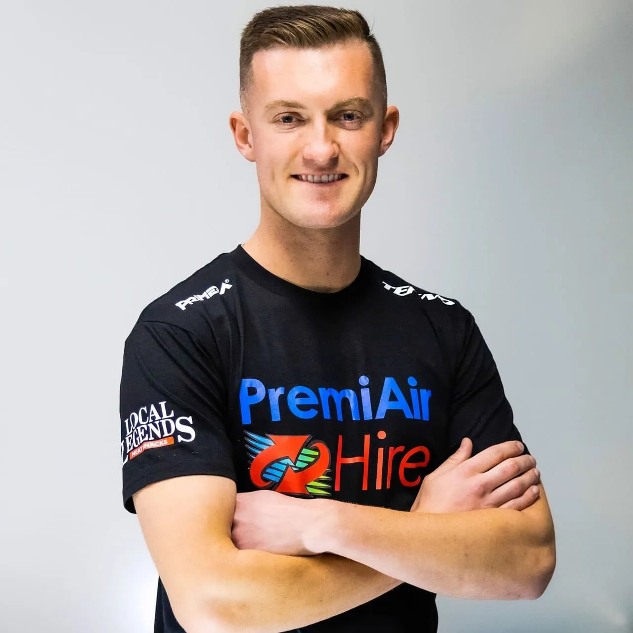 Dylan O’Keeffe smiling with arms folded in a black PremiAir Hire team shirt against a plain grey background
