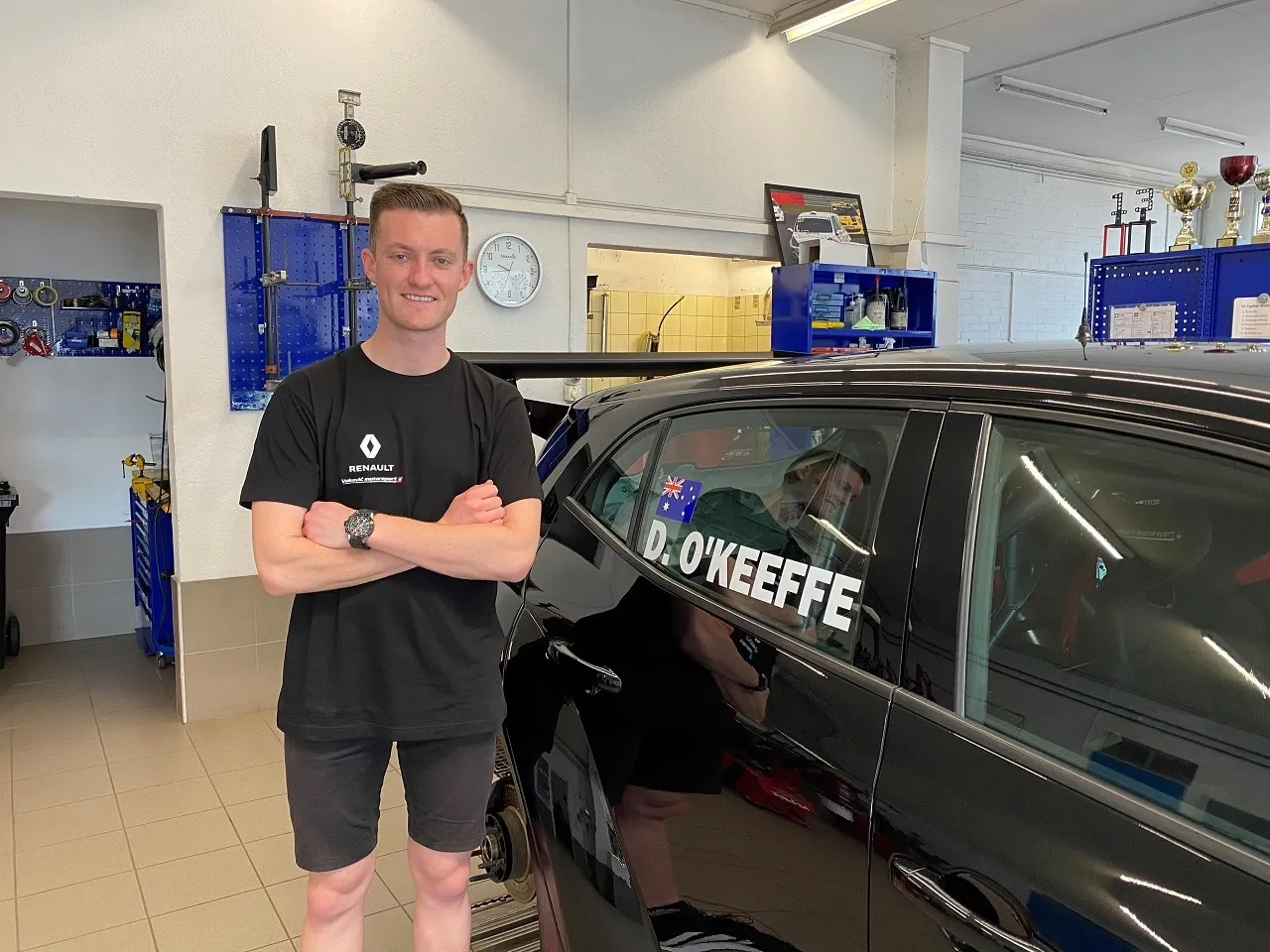 Dylan O’Keeffe smiling with arms folded in a Renault shirt in a workshop, beside a black car with D. O’Keeffe on the window