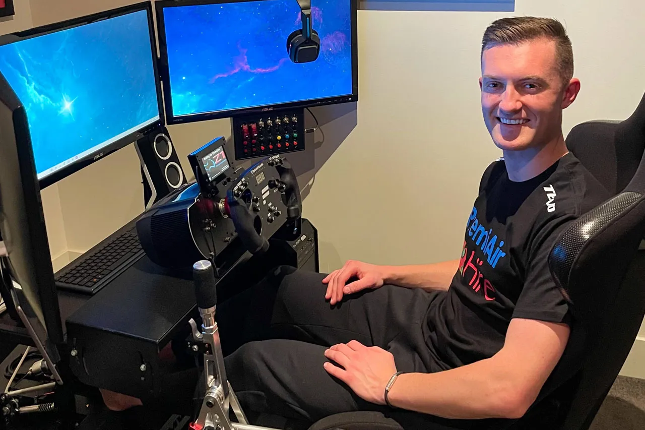 Dylan O’Keeffe smiling in a PremiAir Hire shirt, seated at his sim racing rig with multiple monitors and a wheel