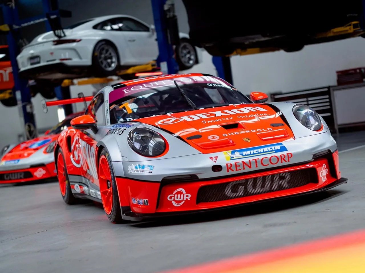Silver and orange Dexion Porsche 911 GT3 Cup car in a workshop with other cars on hoists