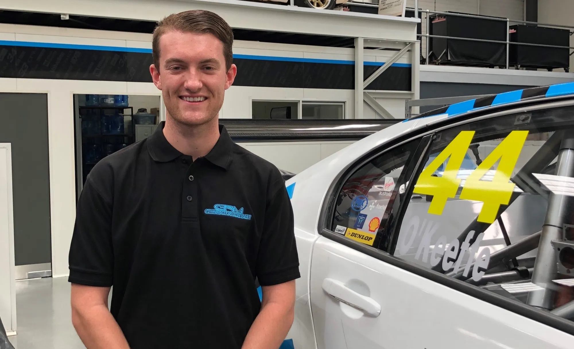 Dylan O’Keeffe smiling in a black Garry Rogers Motorsport polo beside a white race car with #44 and his name on the window
