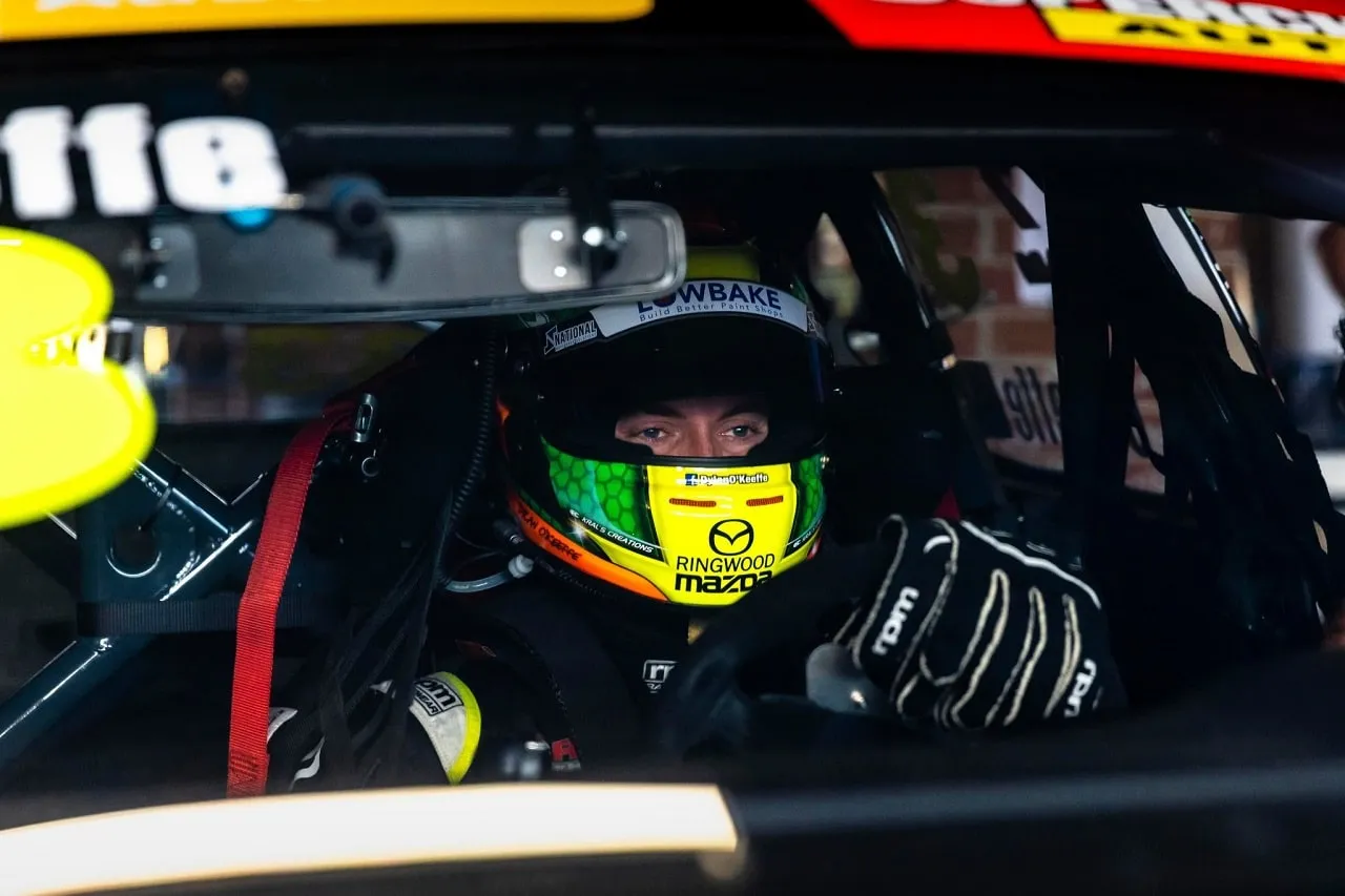 A driver in a green, yellow and black helmet seated in the cockpit of his race car, looking out through the window
