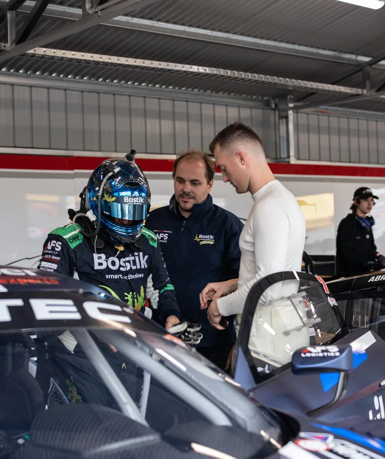 A driver in a Bostik race suit and helmet in the garage speaking with two men beside the Audi R8 GT3 car