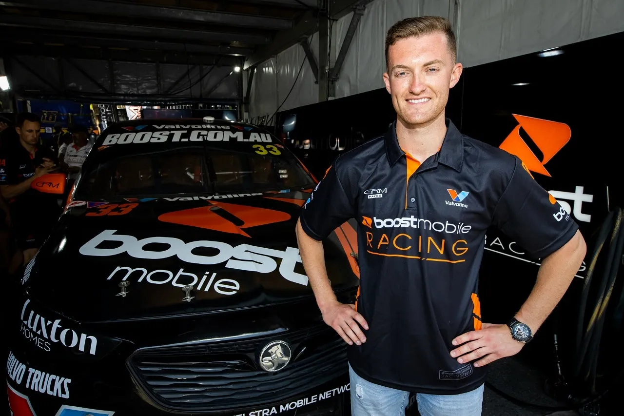 Dylan O’Keeffe smiling in a Boost Mobile Racing shirt beside the black and orange #33 Boost Mobile Commodore in the garage