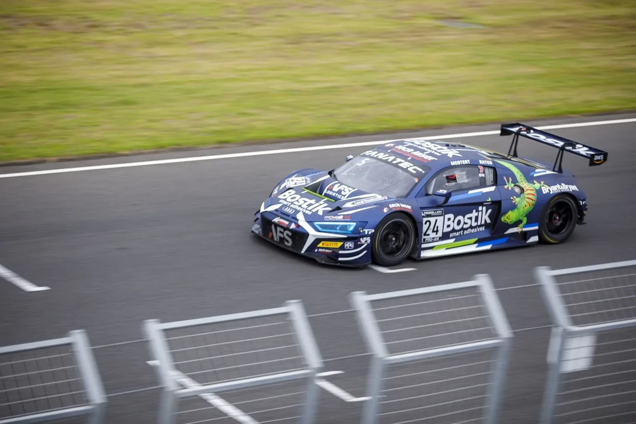The blue #24 Bostik Audi R8 GT3 car on track past a metal fence, seen from a three-quarter angle