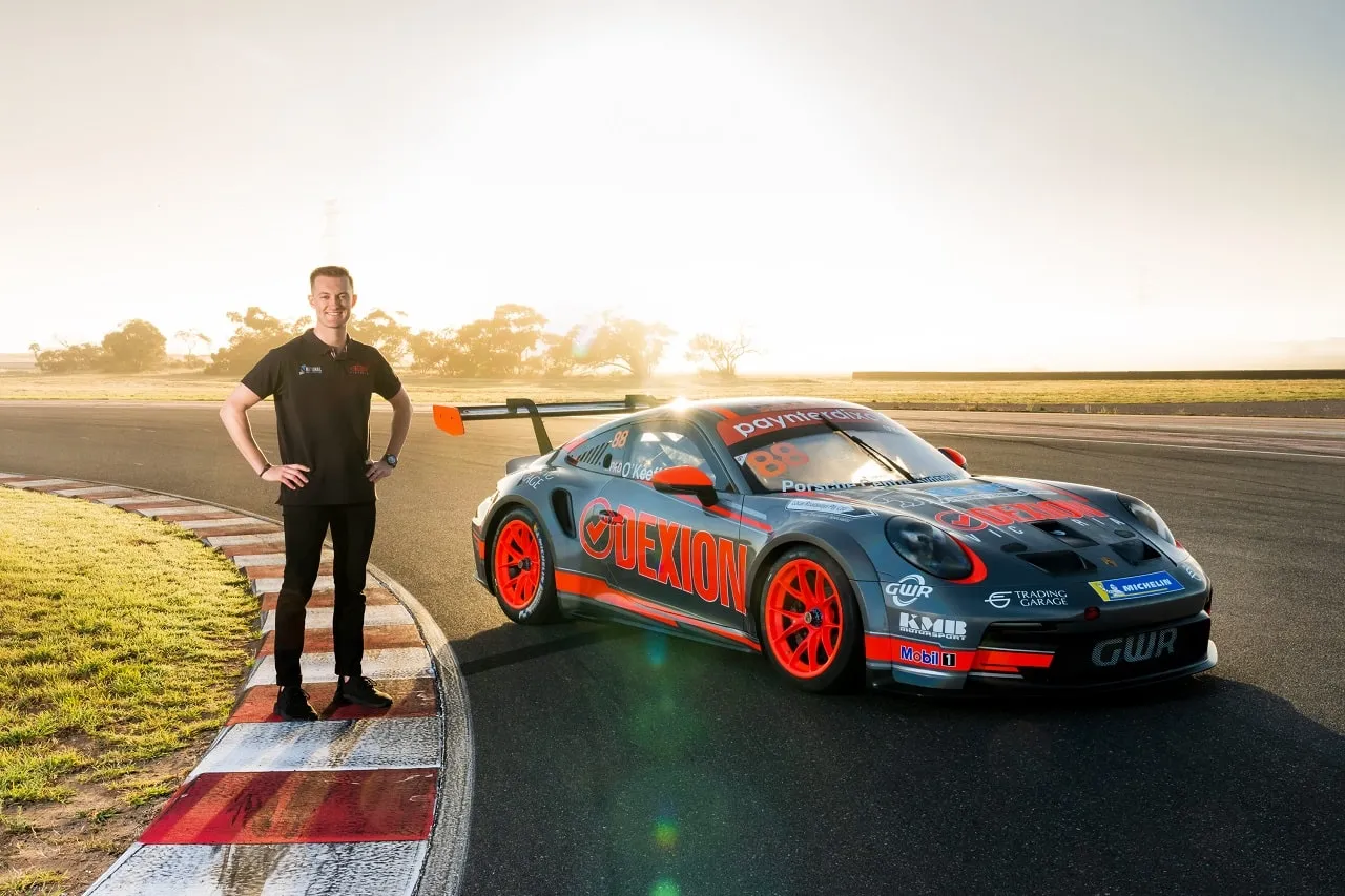 Dylan O’Keeffe standing beside the Dexion Porsche 911 GT3 Cup car on a kerb at sunrise at The Bend