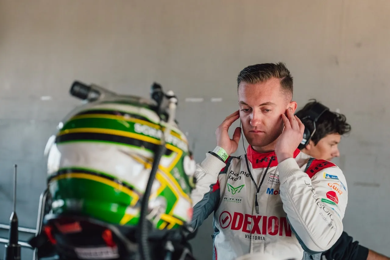 Dylan O’Keeffe in a Dexion Victoria race suit adjusting his headset, with his green and yellow helmet in the foreground