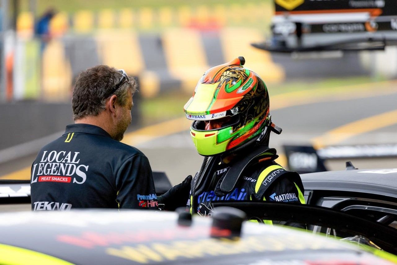 A driver in a green, orange and black helmet talking with a team member in a Local Legends shirt in pit lane