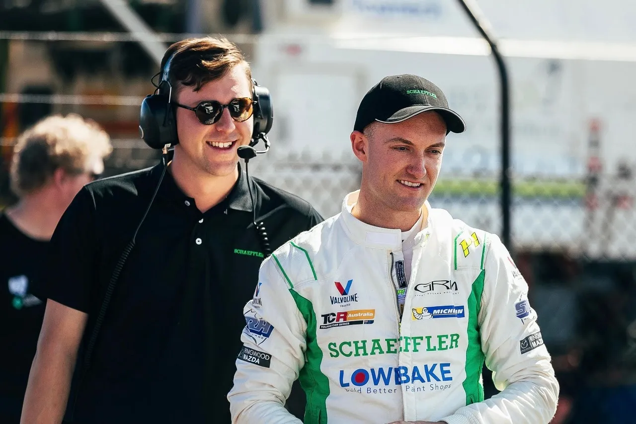 A smiling driver in a white Schaeffler race suit and cap walks beside a man in a headset and sunglasses