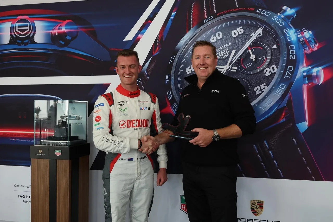 Dylan O’Keeffe in a Dexion race suit shakes hands with a man holding an award in front of a TAG Heuer and Porsche display