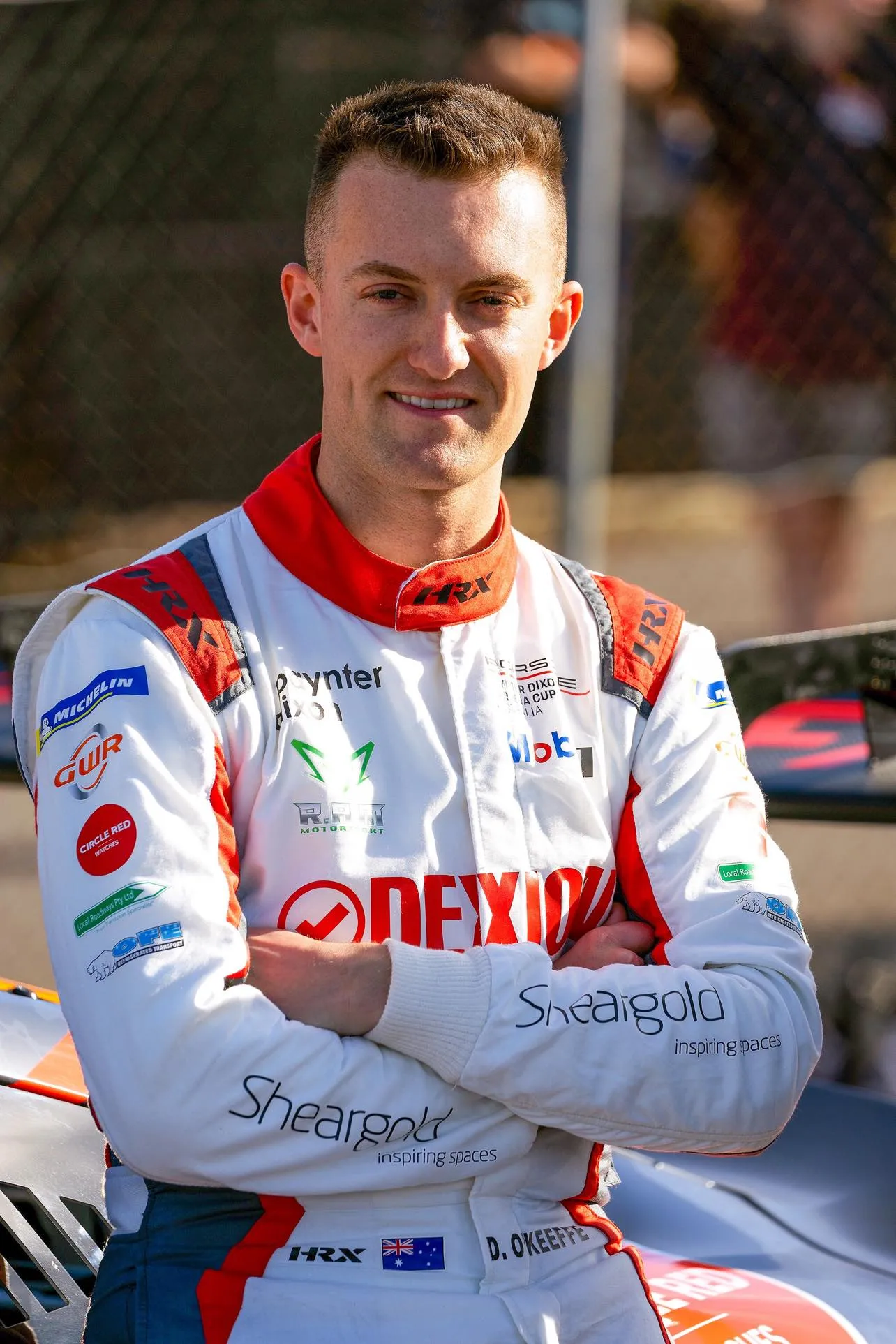 Dylan O’Keeffe in a white Dexion race suit with arms folded, smiling, in front of a Porsche