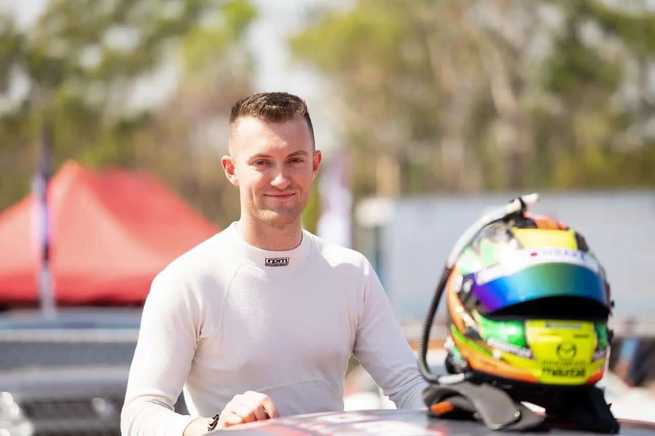 Dylan O’Keeffe in a white race suit smiling beside his helmet in bright sunshine