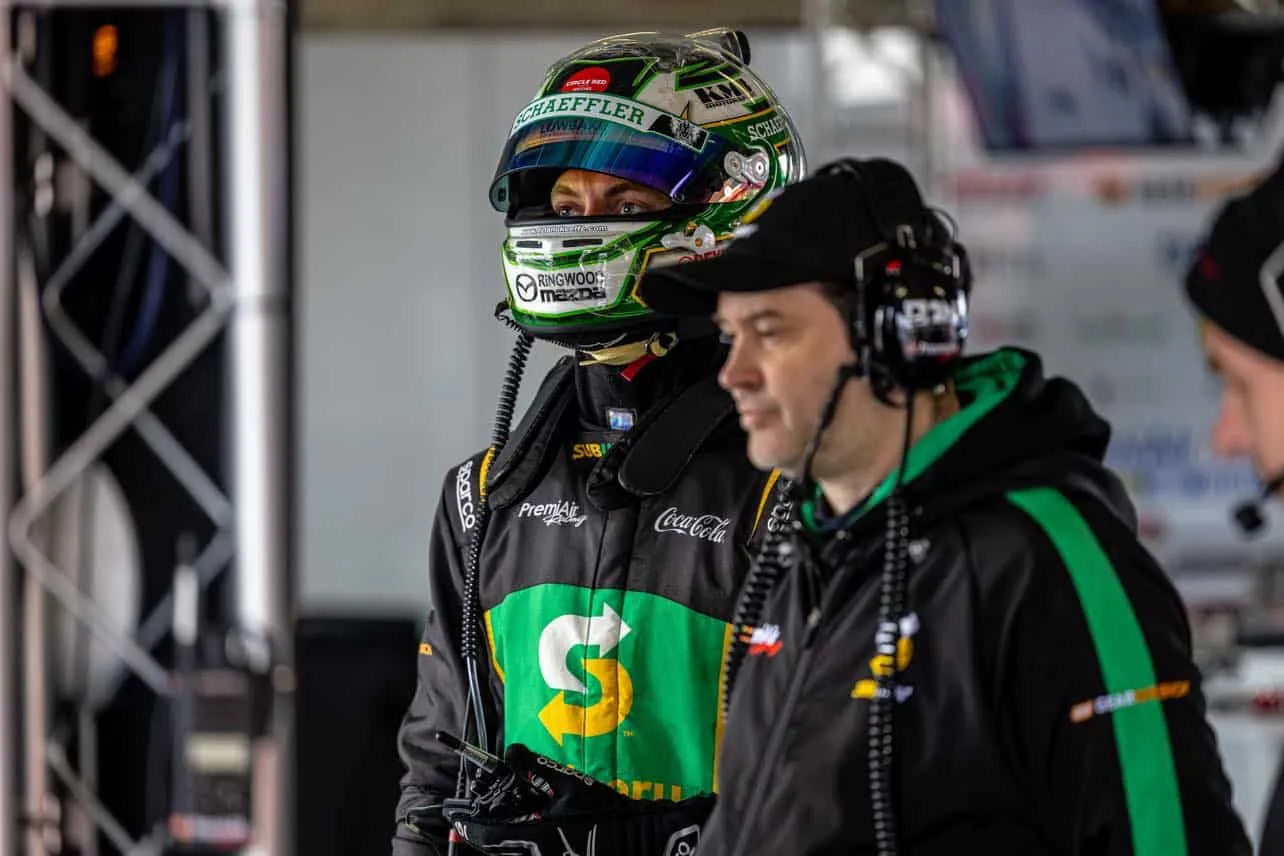 A driver in a green Schaeffler helmet and Subway race jacket stands beside a crew member wearing a headset