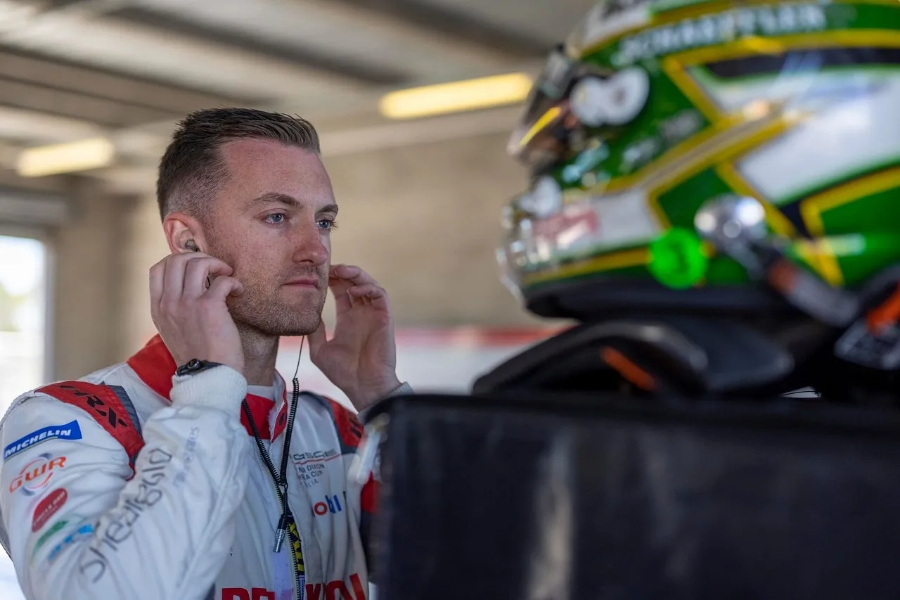 Dylan O’Keeffe in a white race suit adjusting his earpiece in the garage, with his helmet beside him