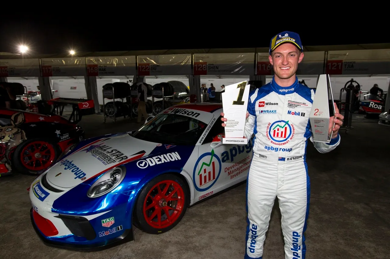 Smiling driver in an APB Group race suit holding two trophies beside a blue Porsche Carrera Cup car at night