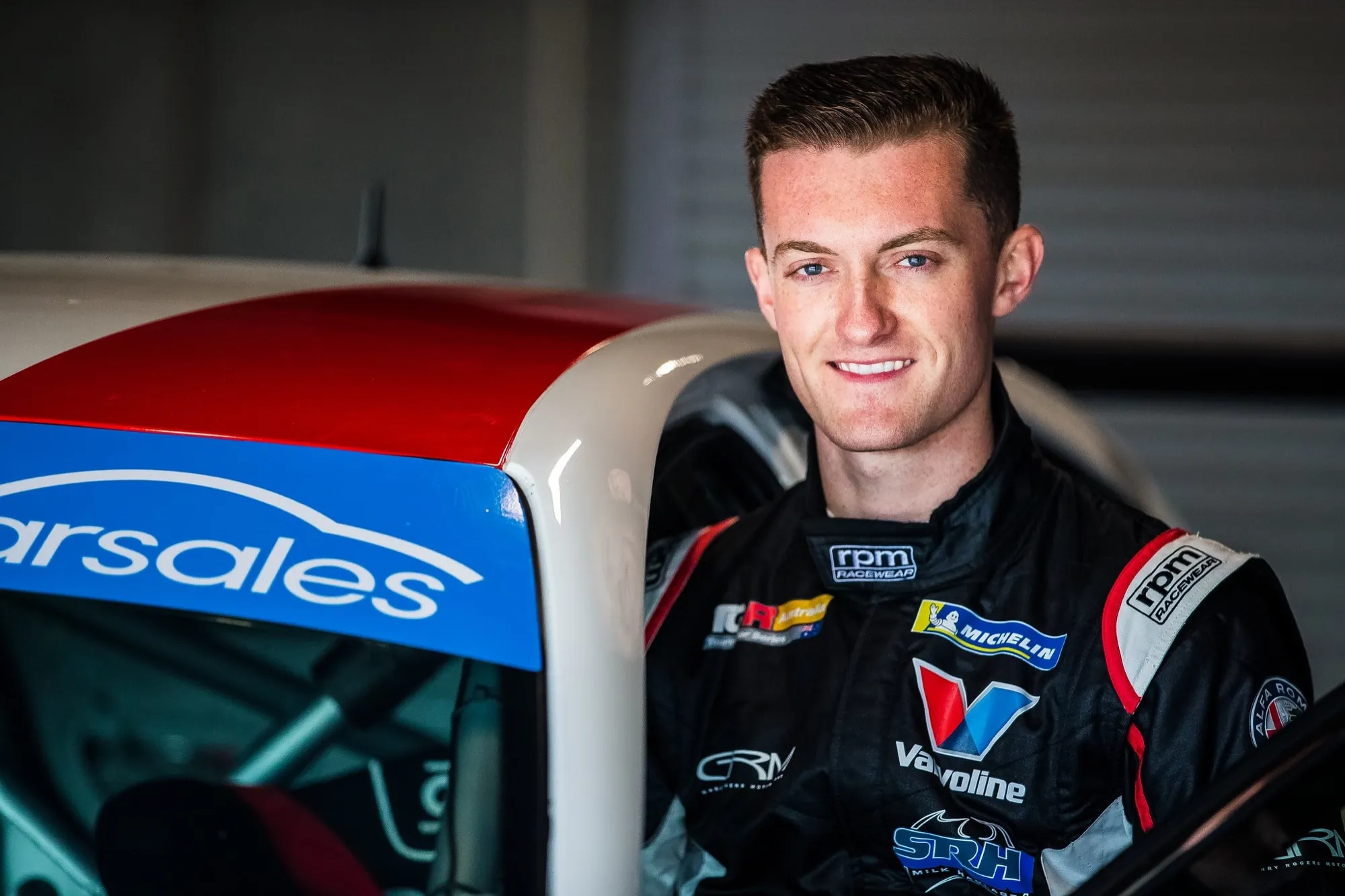 Portrait of Dylan O’Keeffe smiling in his black Valvoline race suit beside his TCR car in the garage