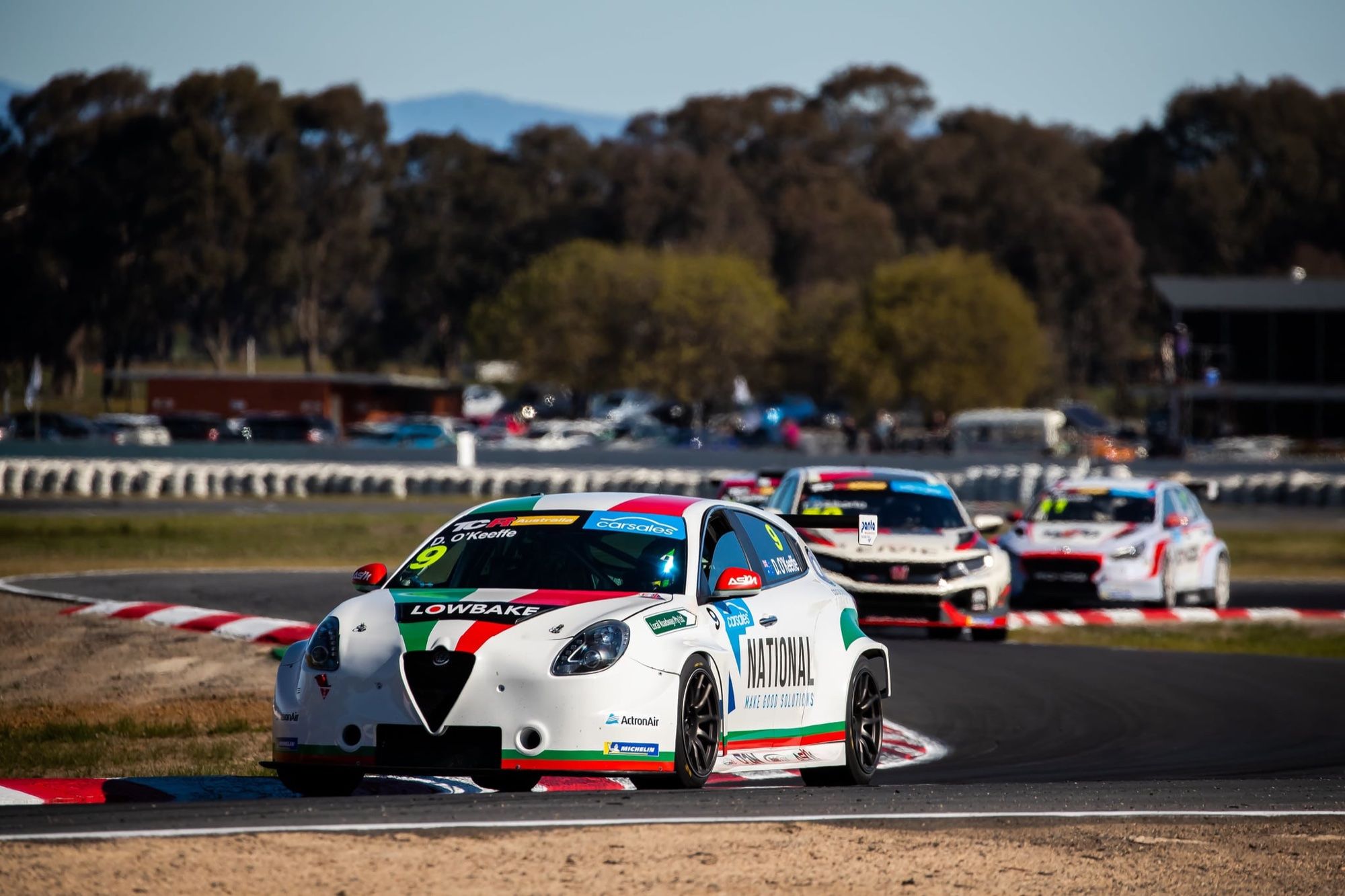 The white #9 Alfa Romeo Giulietta TCR car leading a pack of TCR cars through a corner with trees behind