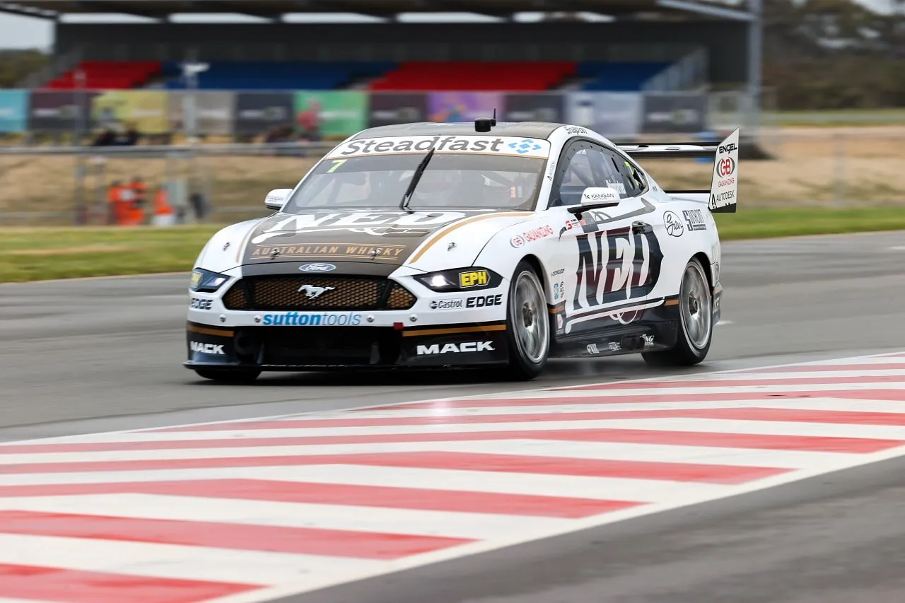 The white NED Whisky Ford Mustang Supercar on track near a red and white kerb, with grandstands behind