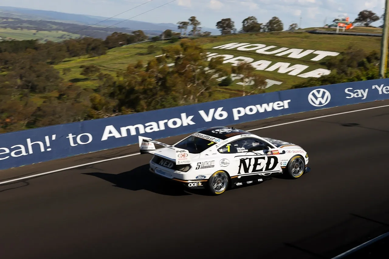 The white NED Whisky Ford Mustang Supercar on track at Mount Panorama beside an Amarok V6 barrier sign