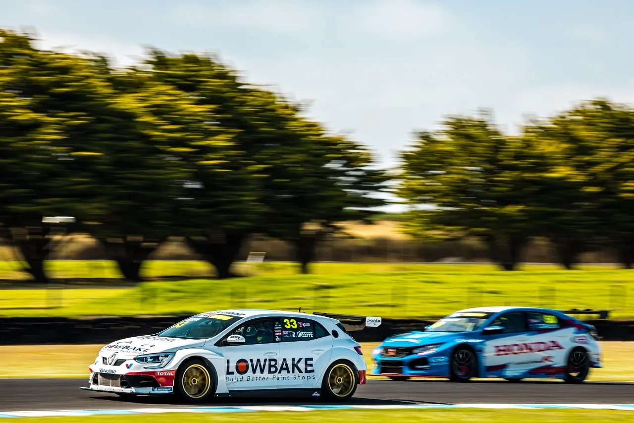 The white #33 Lowbake Renault Megane TCR car on track with a blue Honda Civic TCR car behind, with motion blur in the trees