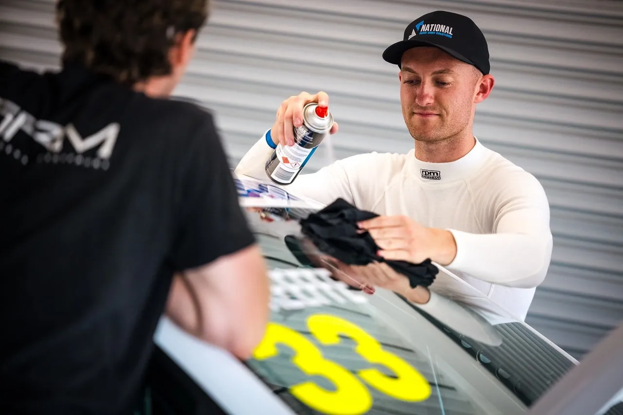 A driver in a National cap and white race suit spraying and wiping the windscreen of the #33 Renault TCR car