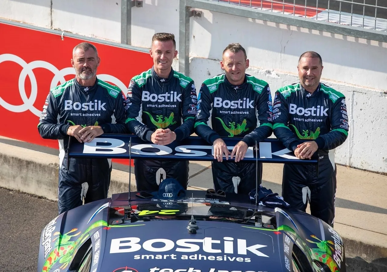 Four drivers in Bostik race suits pose behind the rear wing of the blue Audi R8 GT3 car