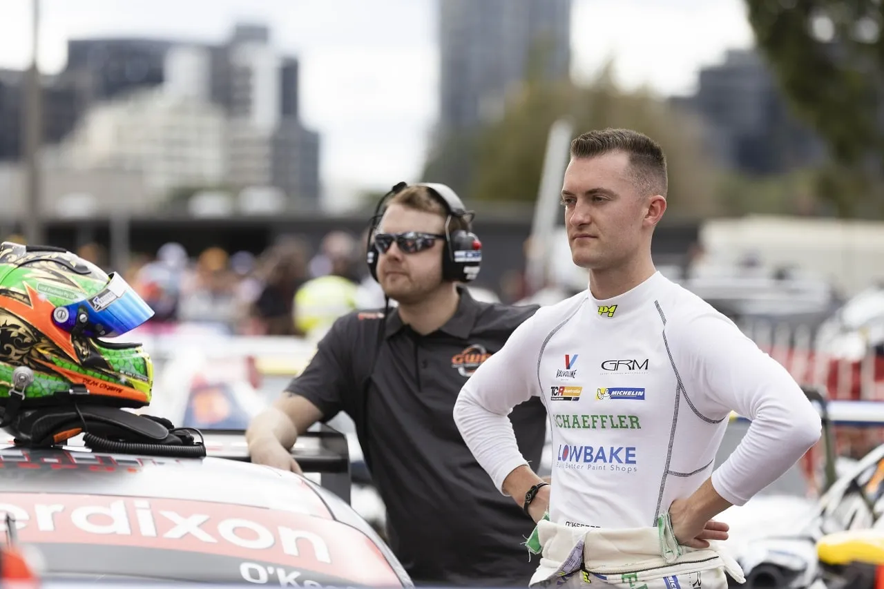 Dylan O’Keeffe in a white Schaeffler race suit stands beside a crew member wearing a headset and sunglasses