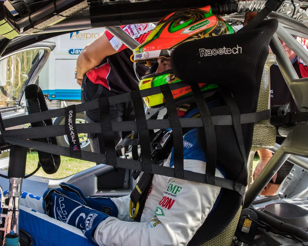 A driver in a green and orange helmet sitting in a race car cockpit behind a window net
