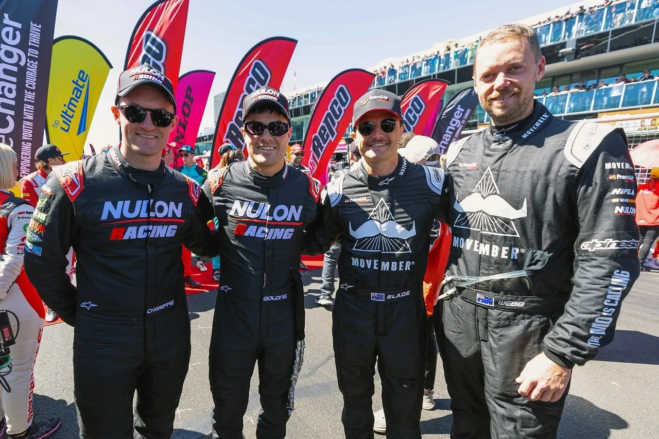 Four drivers in black race suits, two in Nulon Racing suits, posing together in the pit lane at Bathurst