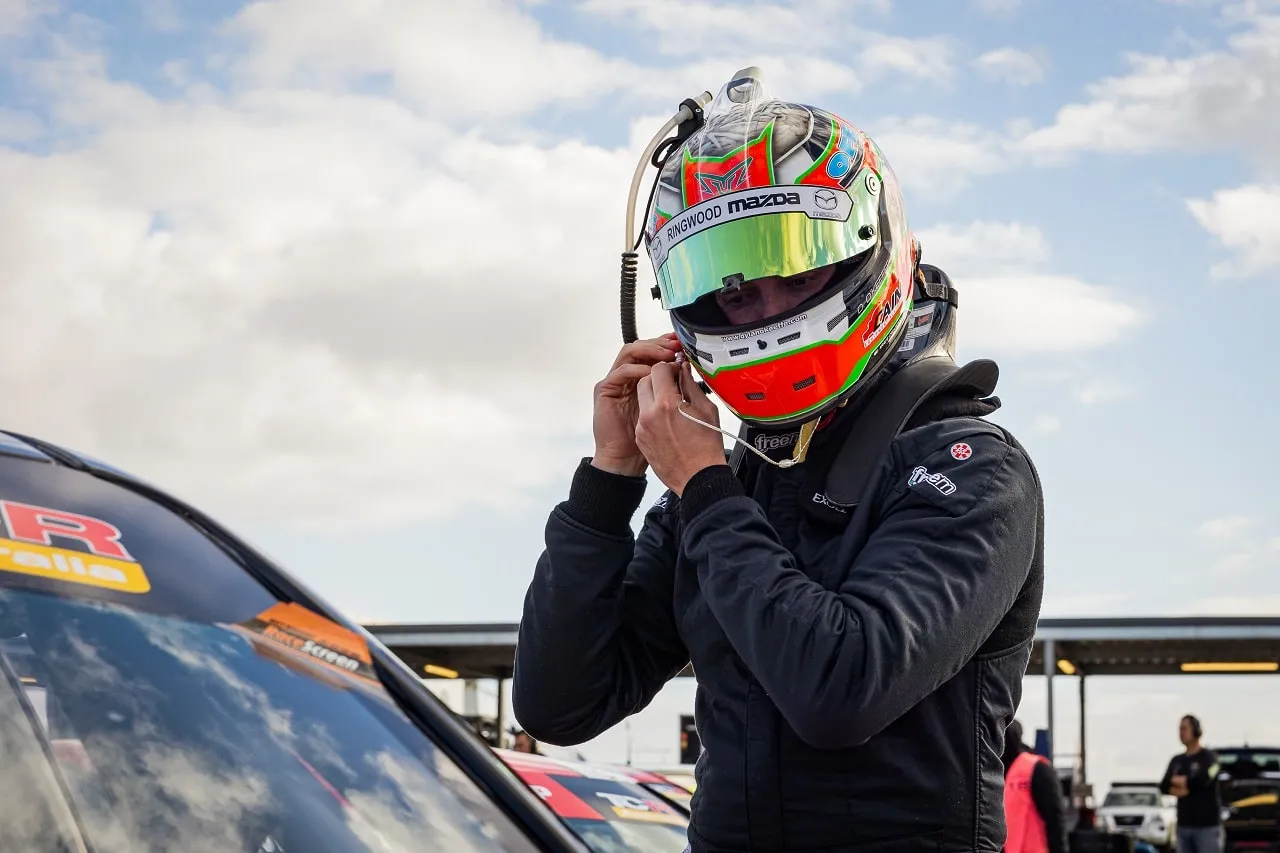 Driver in a helmet and race suit adjusting his chin strap beside a race car
