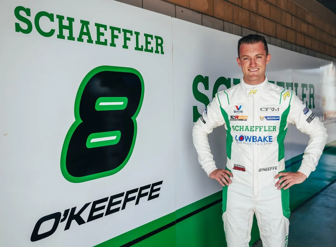Dylan O’Keeffe in a white Schaeffler race suit standing beside a Schaeffler #8 O’Keeffe garage sign