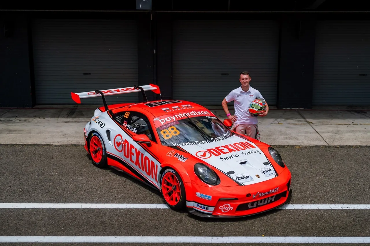 Dylan O’Keeffe holding his helmet beside the orange and white Dexion number 88 Porsche 911 GT3 Cup car in a pit lane