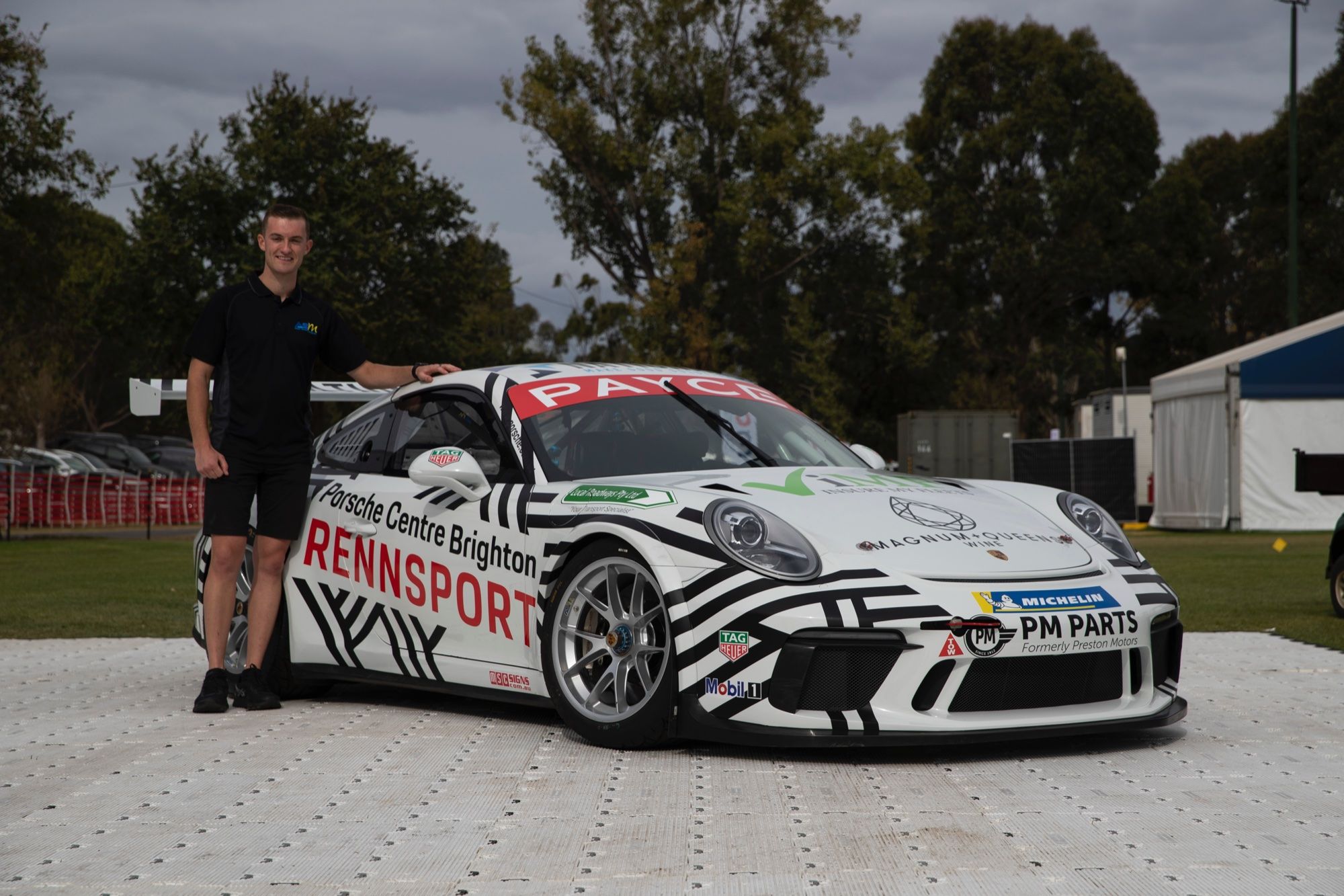 A young man in a black polo standing beside the black and white Porsche Centre Brighton Rennsport Porsche 911 GT3 Cup car