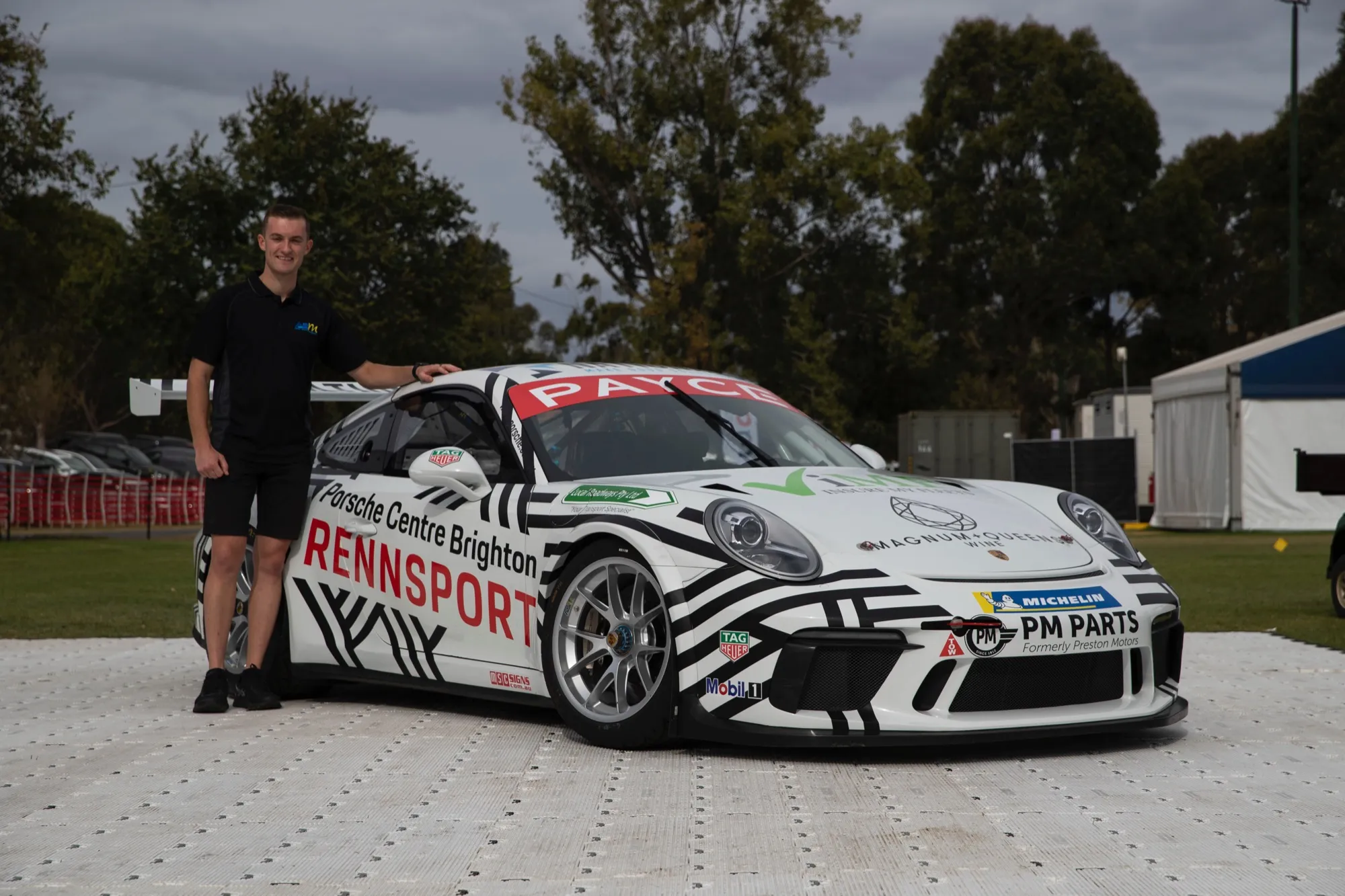 A young man in a black polo standing beside the black and white Porsche Centre Brighton Rennsport Porsche 911 GT3 Cup car
