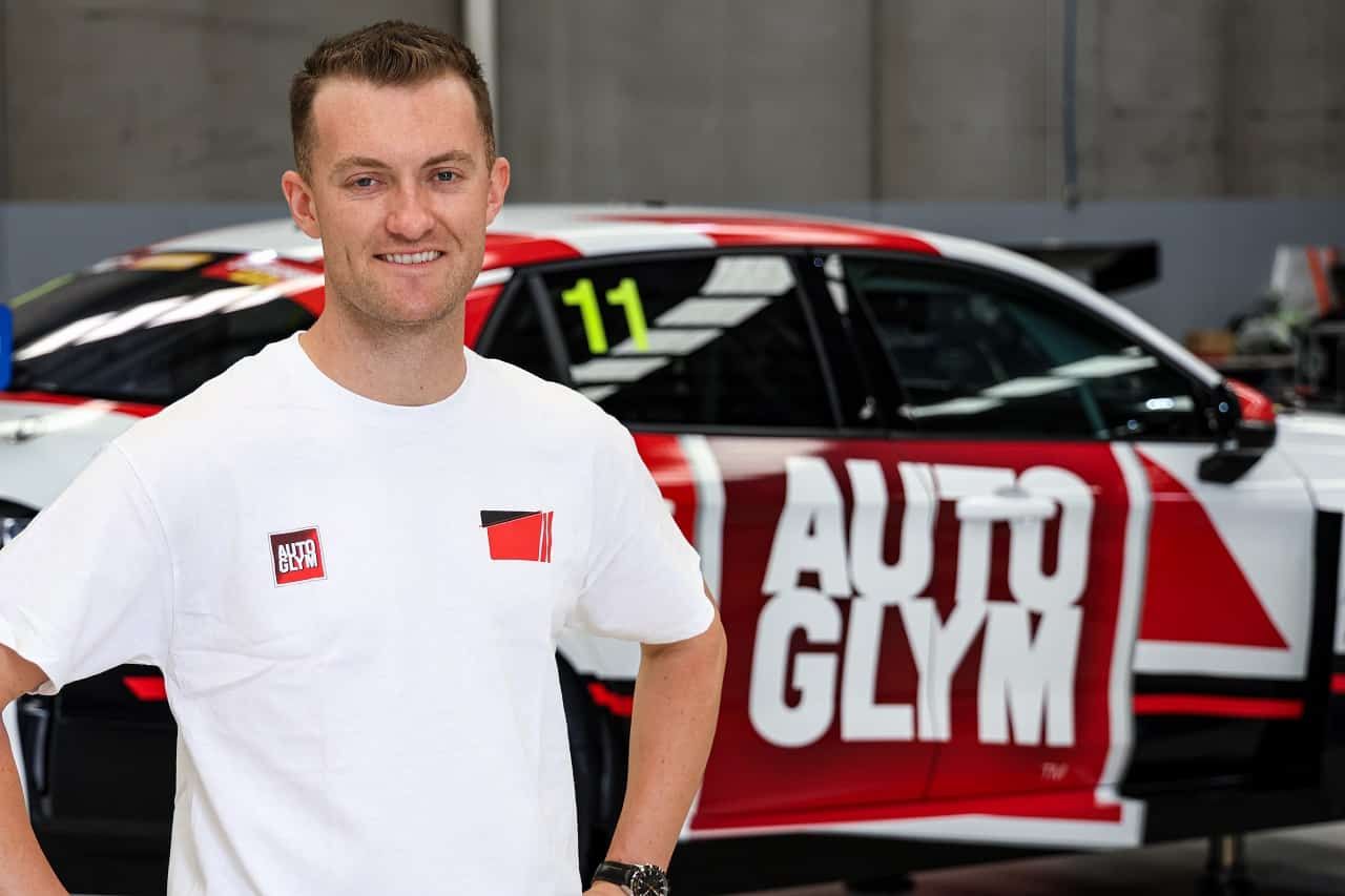 Dylan O’Keeffe smiling in a white T-shirt in front of the Autoglym-liveried Lynk & Co TCR car, number 11