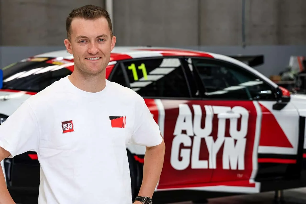 Dylan O’Keeffe smiling in a white T-shirt in front of the Autoglym-liveried Lynk & Co TCR car, number 11