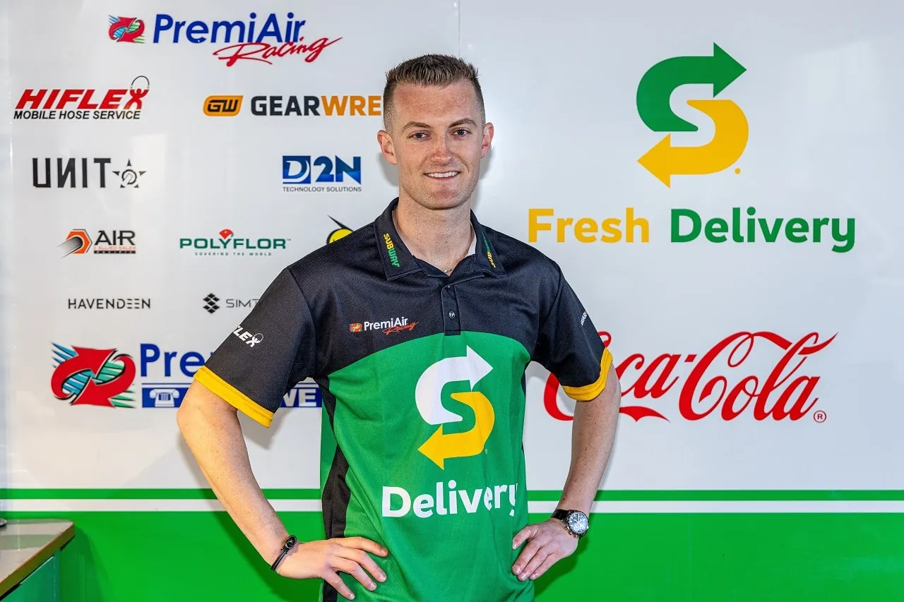 Dylan O’Keeffe smiling in a green Subway Delivery PremiAir Racing shirt in front of a sponsor wall