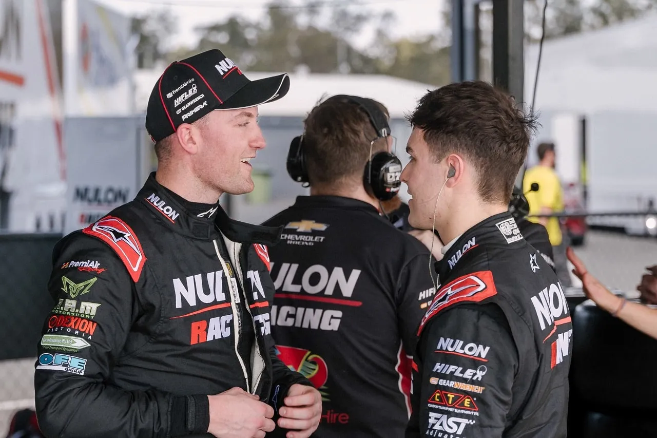 A driver in Nulon Racing overalls and a cap chatting with a younger driver in the garage