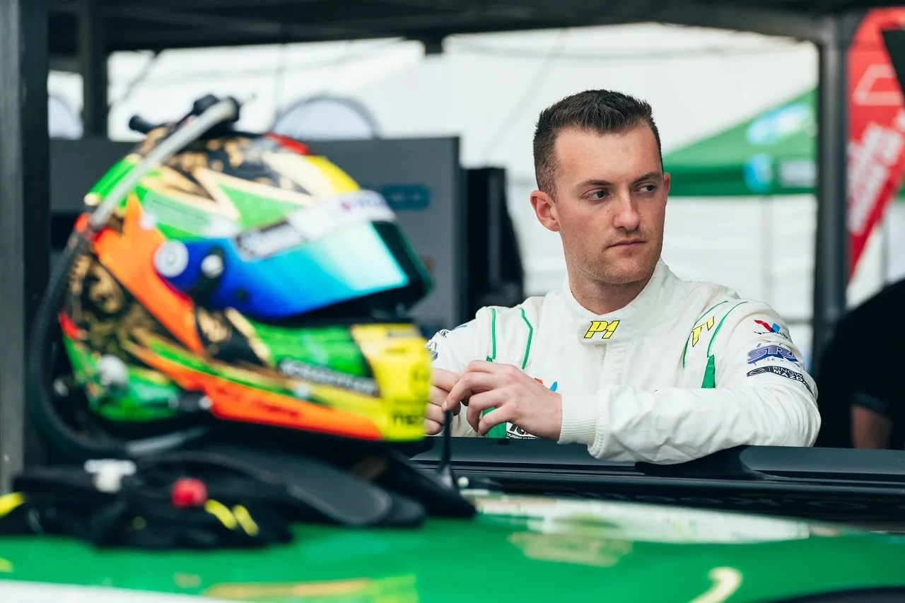 Dylan O’Keeffe in a white race suit looks off to the side beside his green and orange helmet