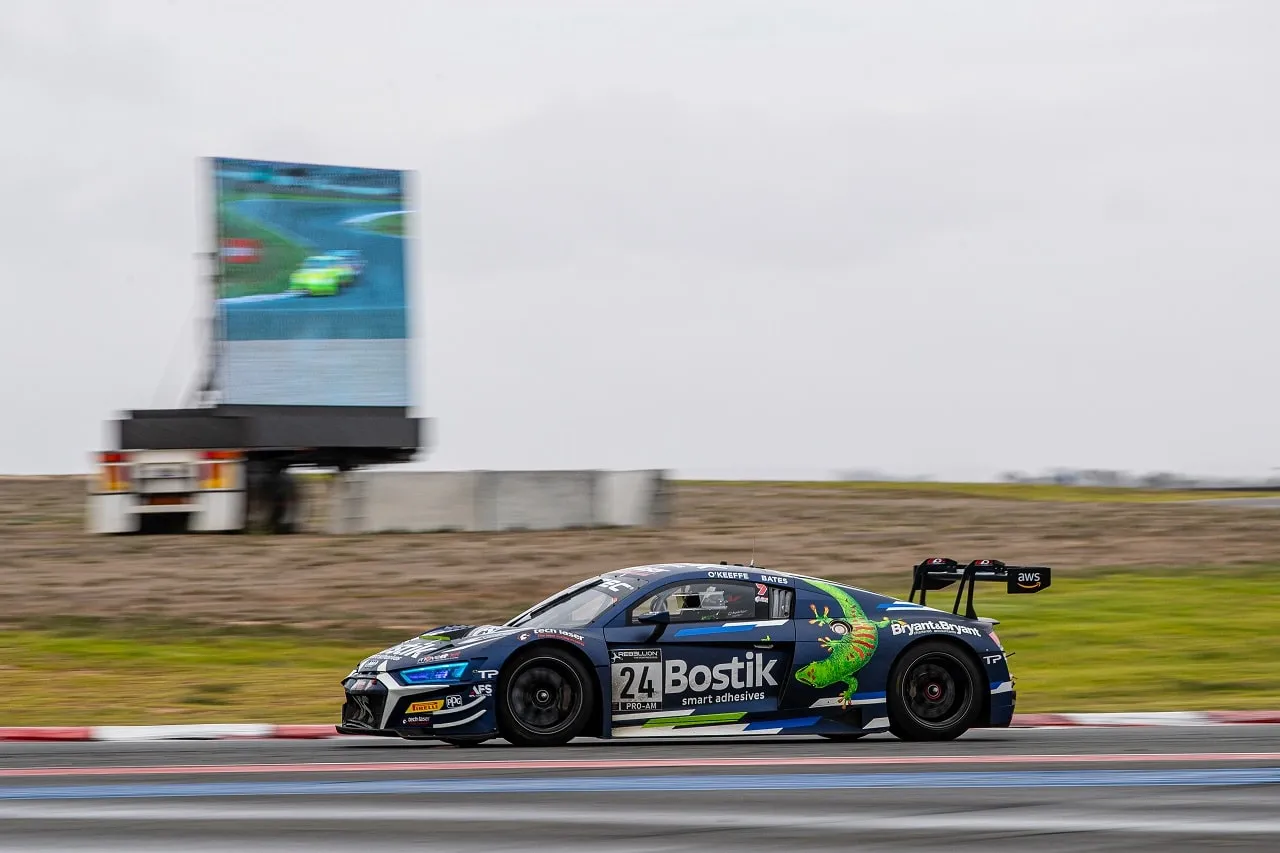 The blue #24 Bostik Audi R8 GT3 car on track at The Bend under an overcast sky, with a video screen behind