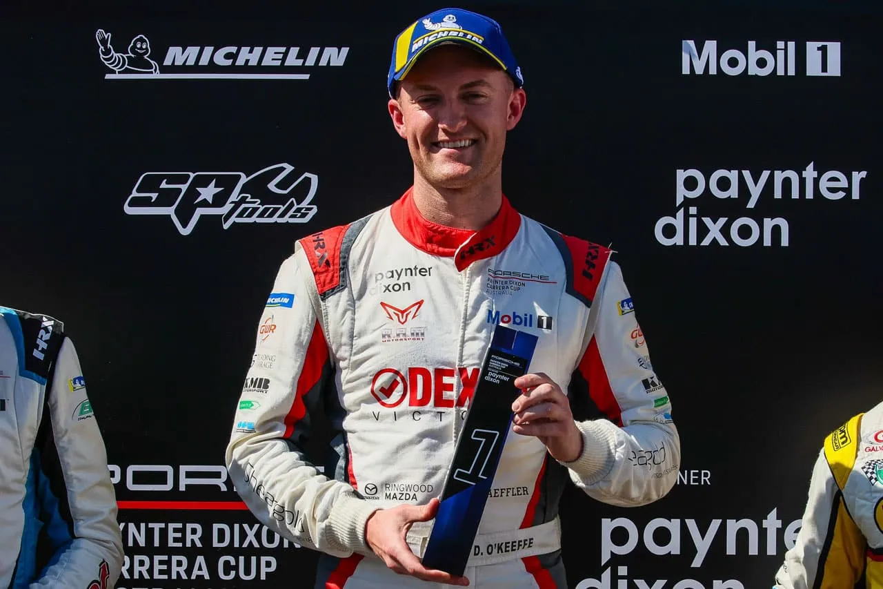 A smiling driver in a Dexion Victoria race suit and Michelin cap holding a number 1 trophy on the podium