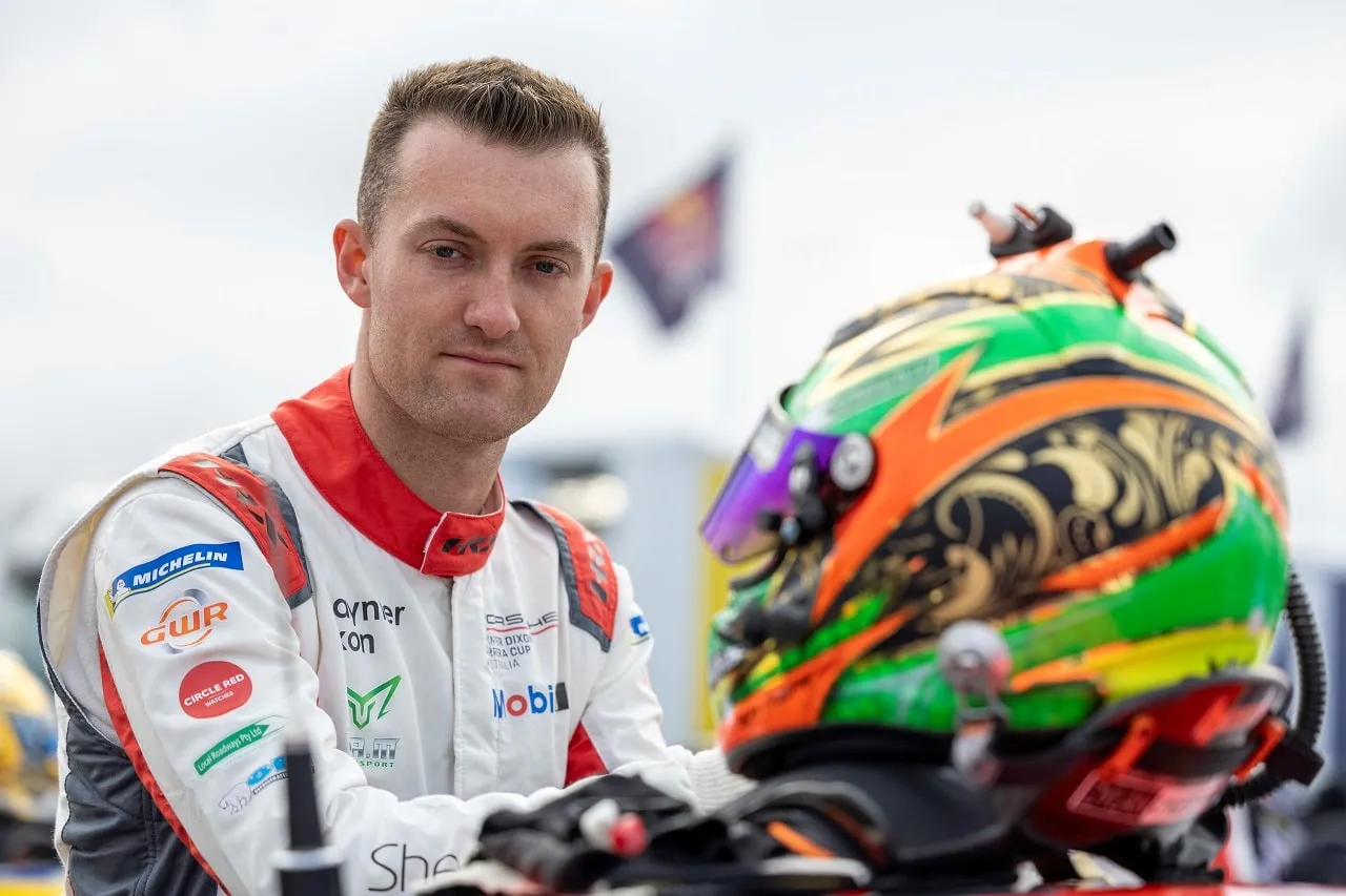 A driver in a white GWR Australia race suit stands beside his green and orange helmet
