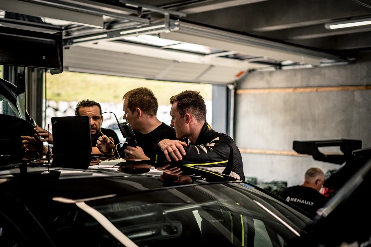 Team members and a driver in a garage leaning over a race car’s roof and working at a laptop
