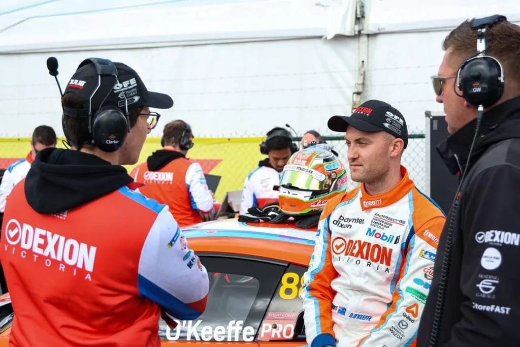 Driver in a Dexion race suit talks with headset-wearing team members beside the number 88 Porsche