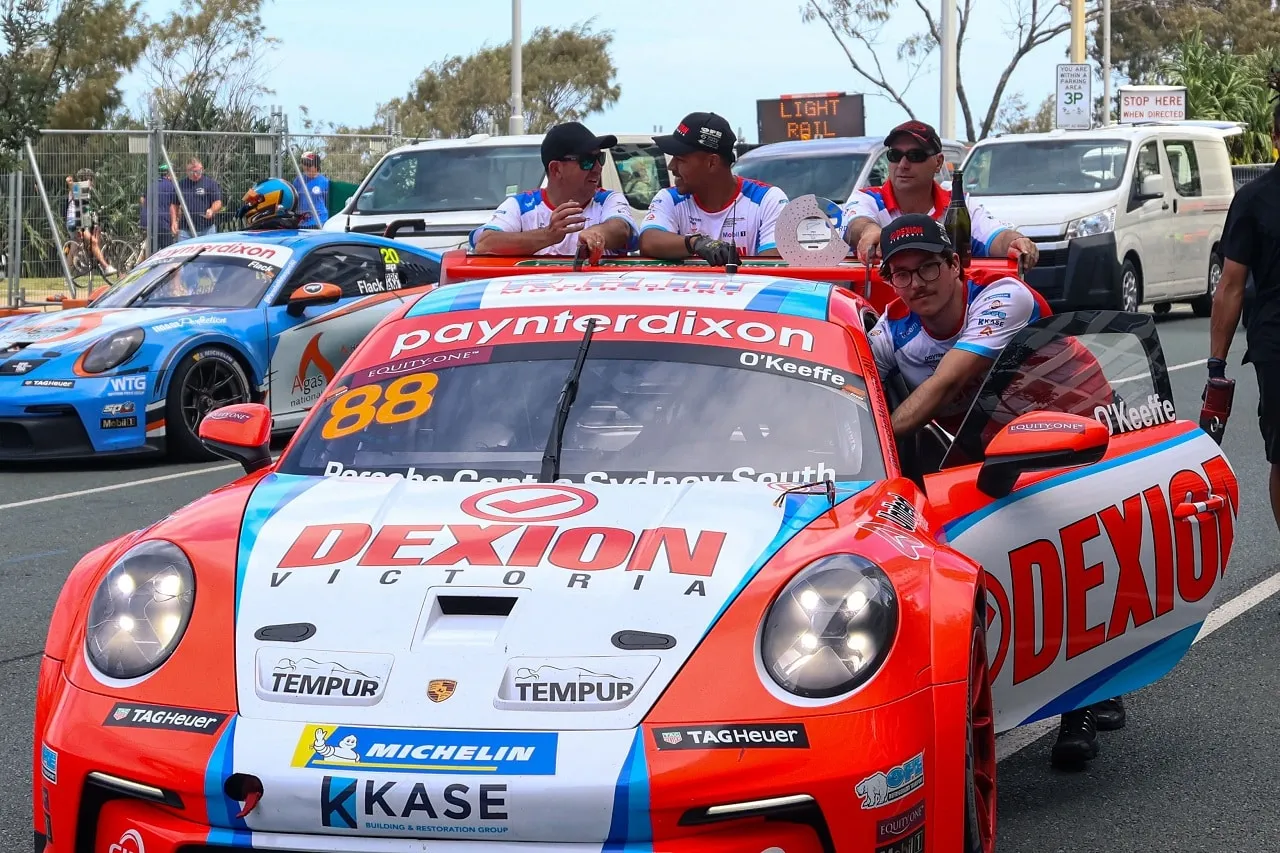 Crew members ride on the front of the number 88 Dexion Porsche Carrera Cup car as it is moved along a street