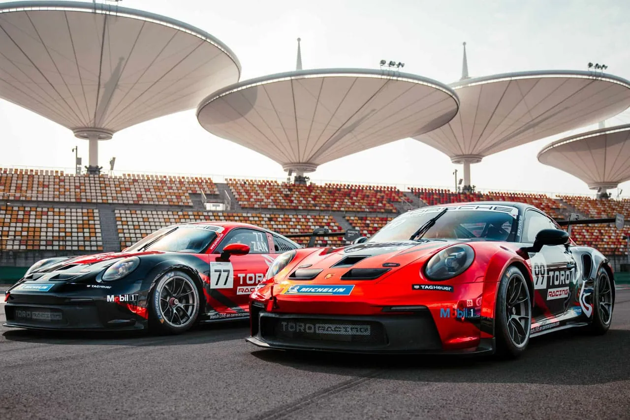 Two Toro Racing Porsche 911 GT3 Cup cars, one black and one red, parked in front of a grandstand with canopies