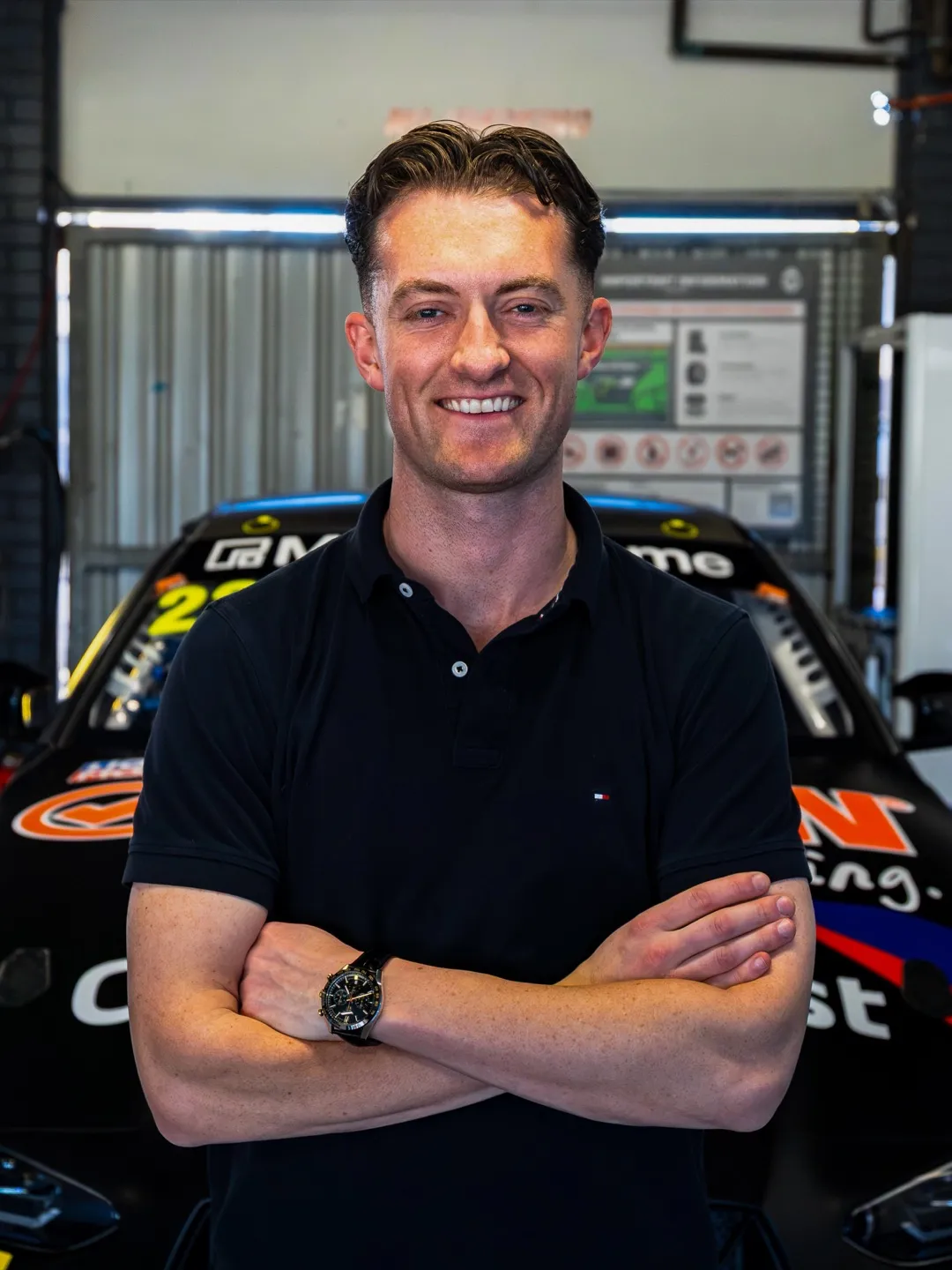 A smiling man in a dark team shirt in a garage, with a Mercedes-AMG car behind him