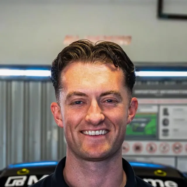A smiling man in a dark team shirt in a garage, with a Mercedes-AMG car behind him
