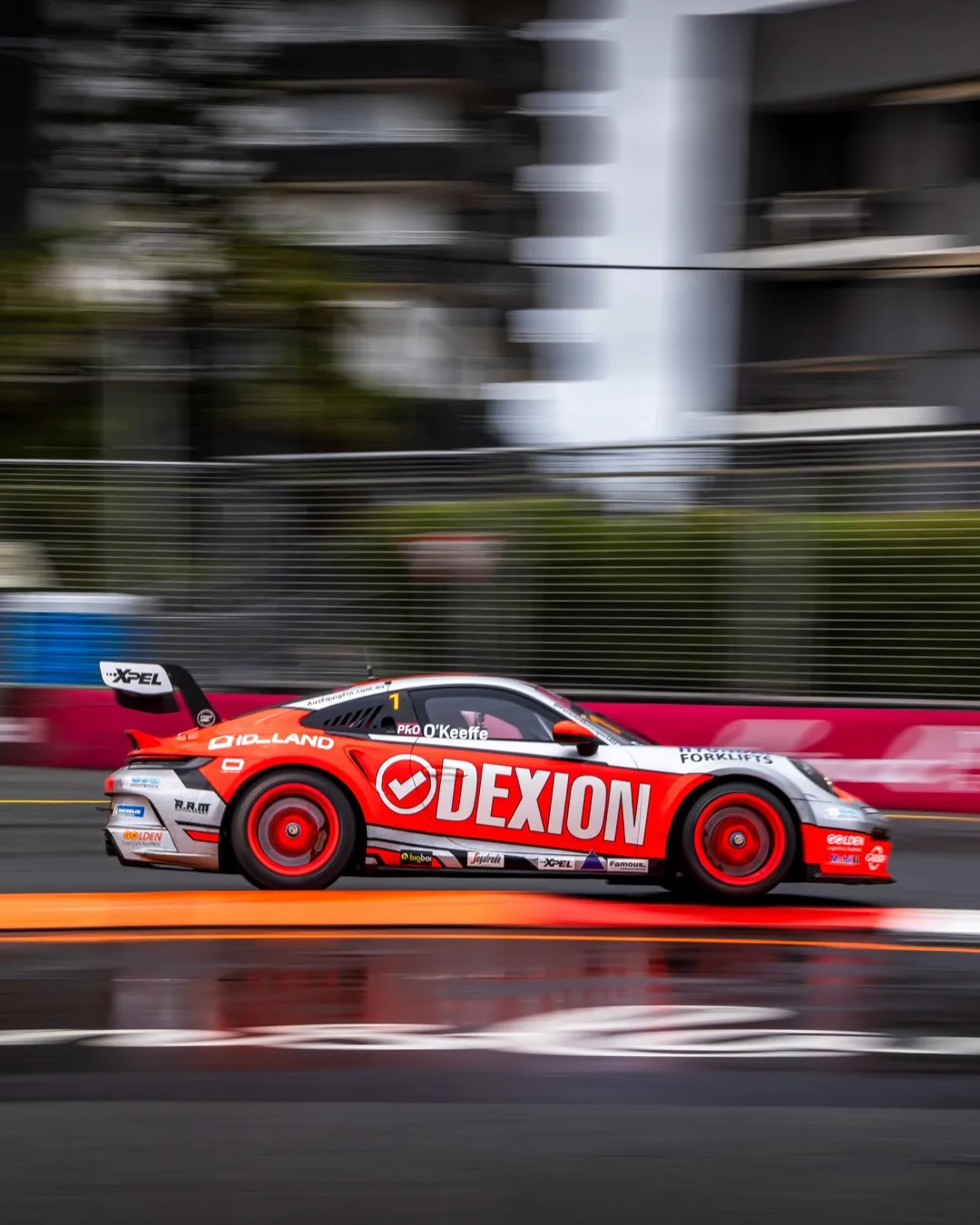 The red Dexion Porsche 911 Carrera Cup car on the Gold Coast street circuit, blurred apartment towers and catch fencing streaking past behind it